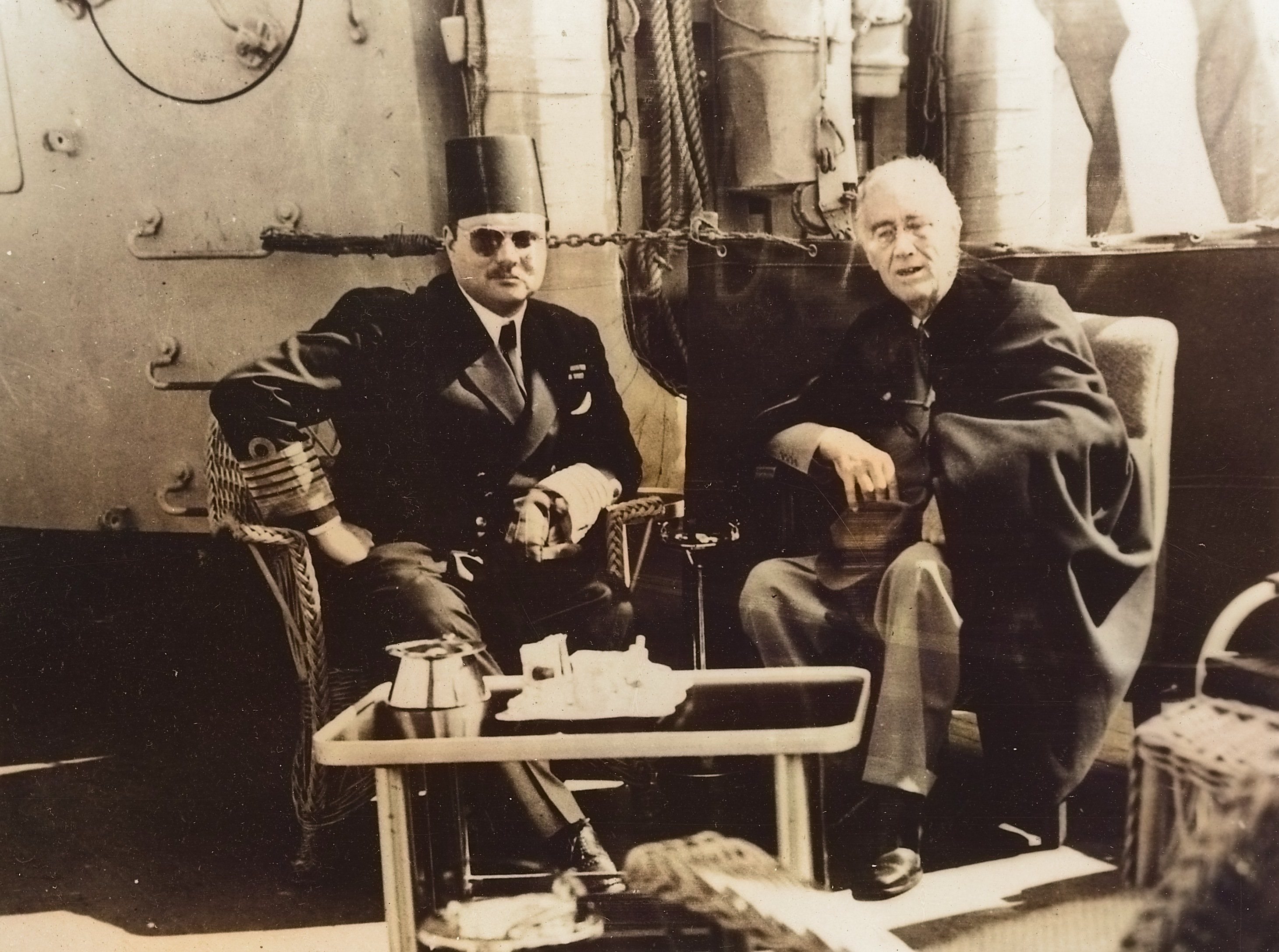 Franklin D. Roosevelt and King Farouk of Egypt seated together outdoors, with naval officers standing behind them.
