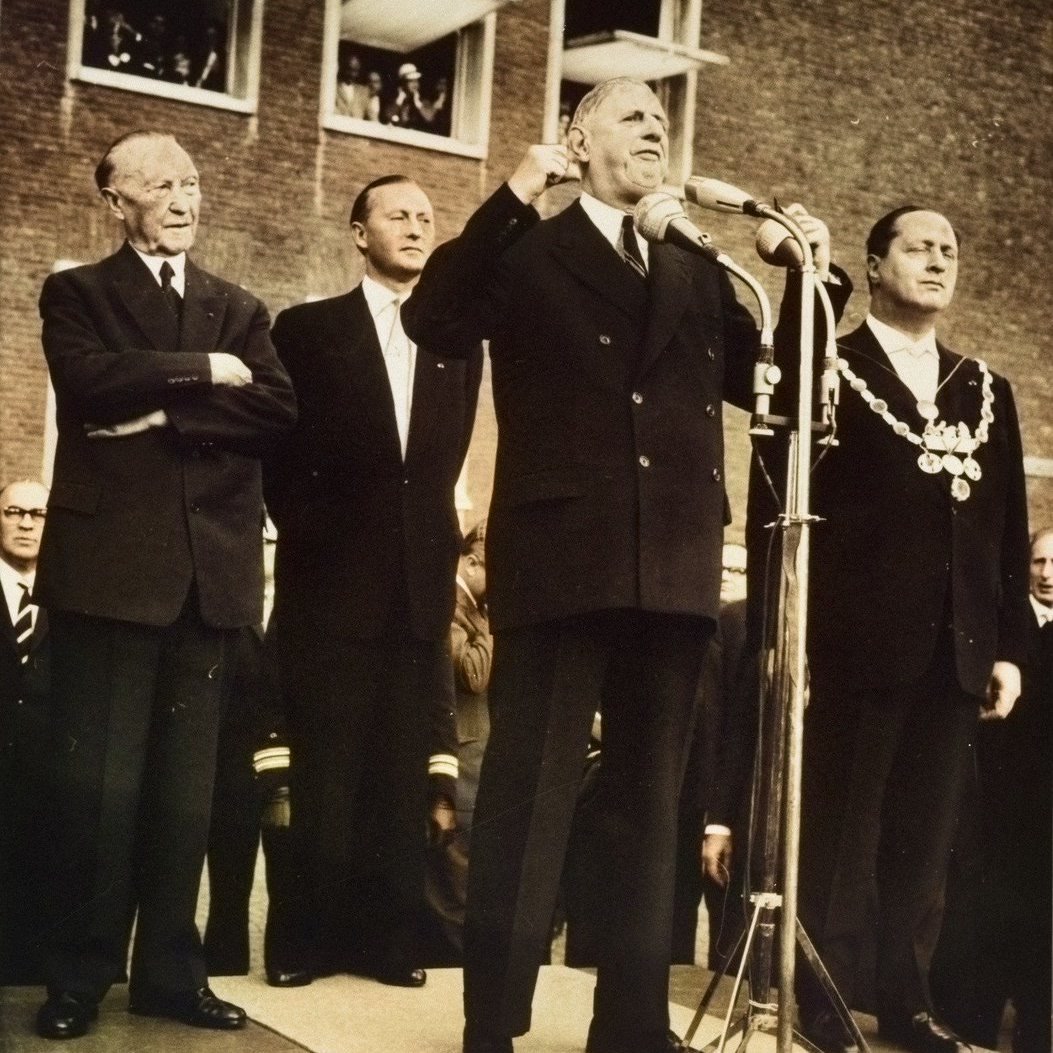 Charles de Gaulle in a dark overcoat speaking into a cluster of microphones, with a crowd behind him.