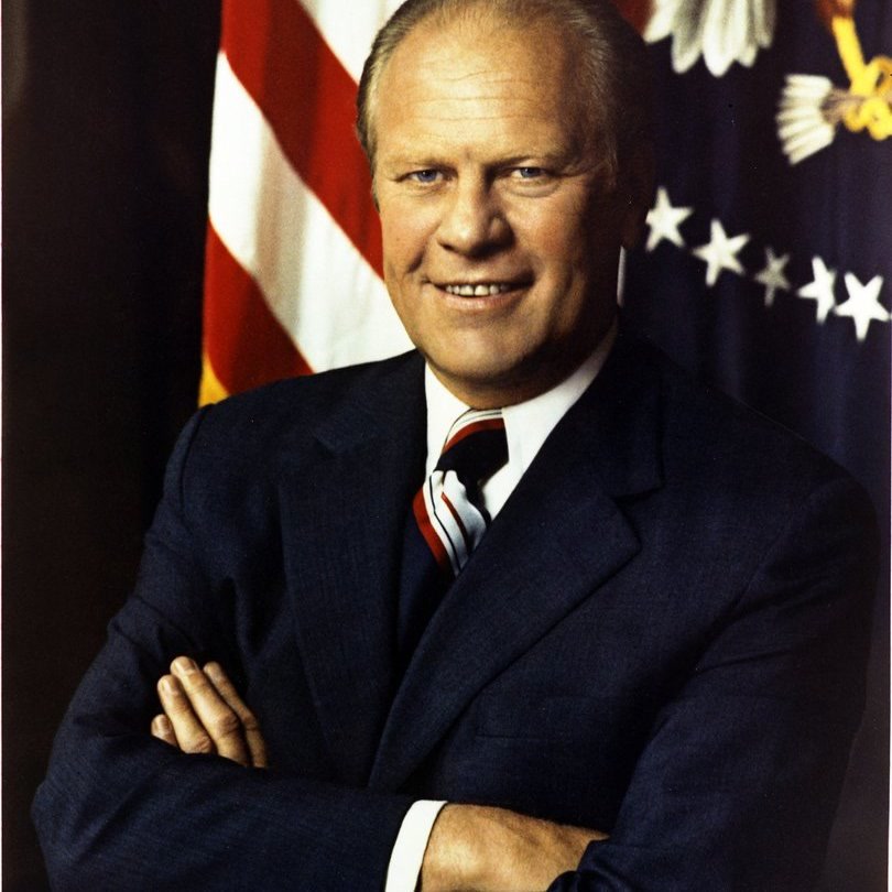 Color portrait of Gerald Ford in a dark suit and patterned tie, standing before an American flag.