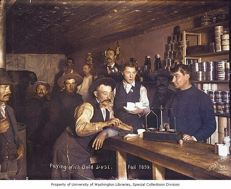 Colorized 1899 photograph of a man in a shop pouring gold dust from a pouch onto a brass scale while another man watches.