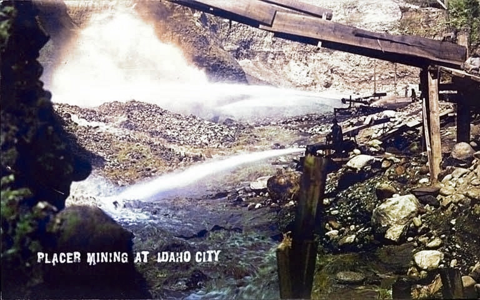Colorized photograph of placer miners directing jets of water from a large metal nozzle into a gravel bank at Idaho City.