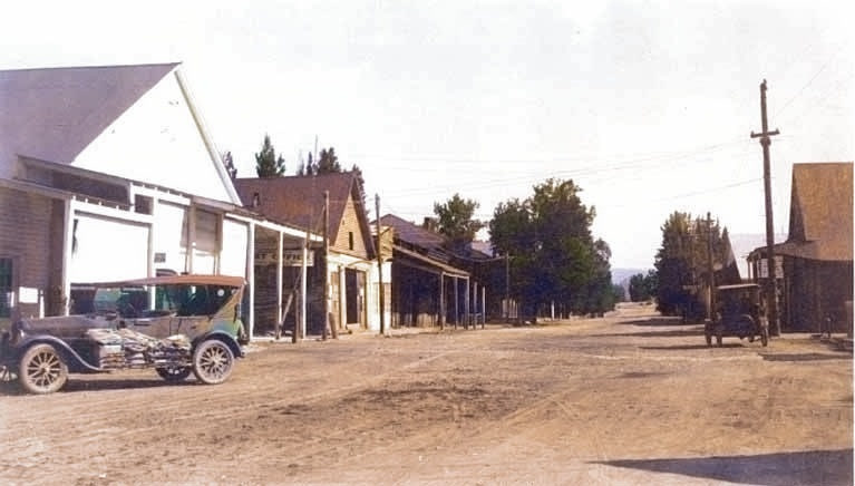 Colorized photograph of a wide dirt main street in Idaho City lined with wooden false-front buildings and a few horses.